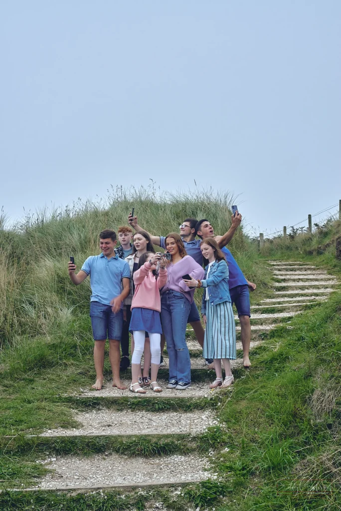 Six people stand on outdoor steps surrounded by grass, holding up mobile phones and taking selfies under a cloudy sky, capturing candid moments as if guided by a family photographer.Family photographer