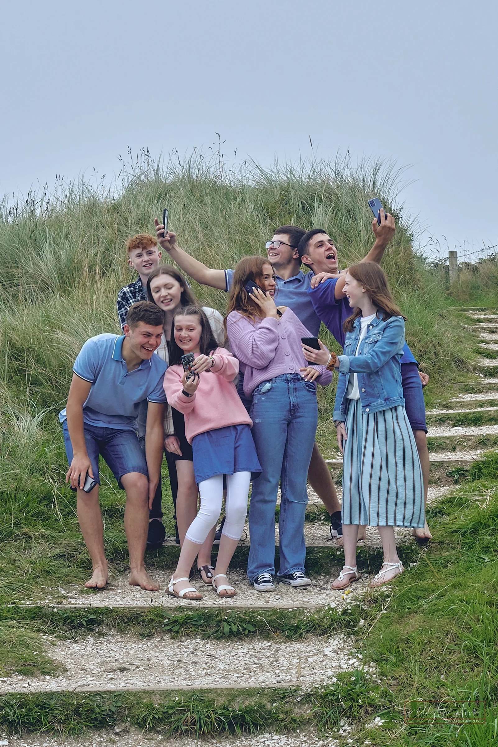 A group of young people stand on outdoor steps, posing for a selfie with mobiles, surrounded by grass and cloudy skies—capturing memories just as a family photographer would.Family photographer