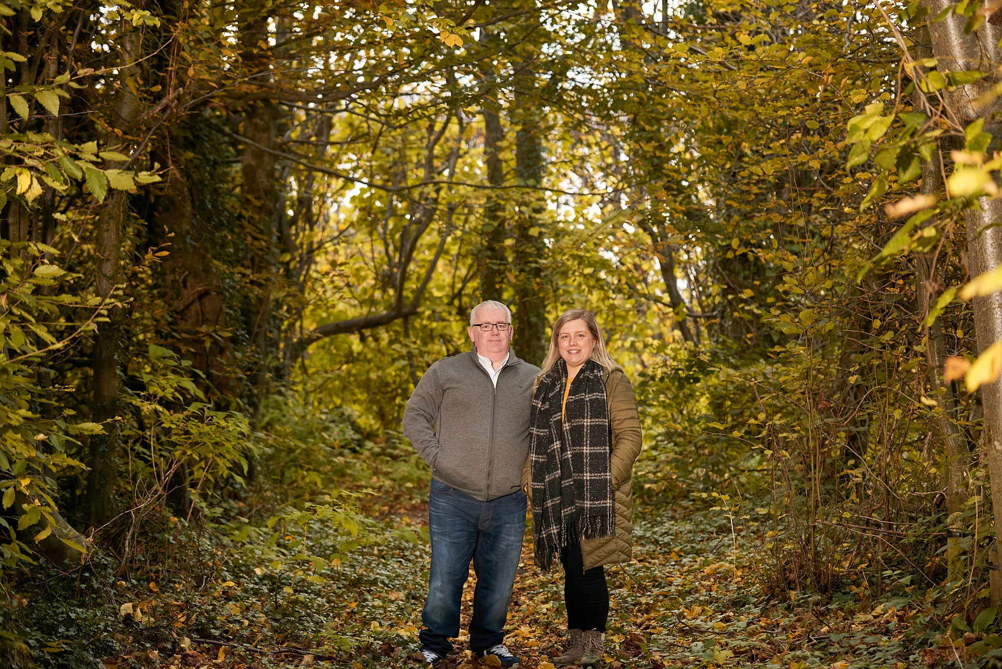 A man and woman stand side by side on a leaf-covered path in a wooded area with yellow and green foliage, capturing a moment ideal for a family photographer.Family photographer
