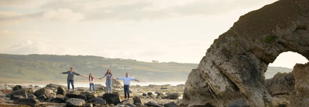 Four people stand holding hands on rocky terrain near a large rock formation with a hole, whilst hills and houses are visible in the background under a cloudy sky—an ideal scene for any family photographer to capture memorable moments.Family photographer