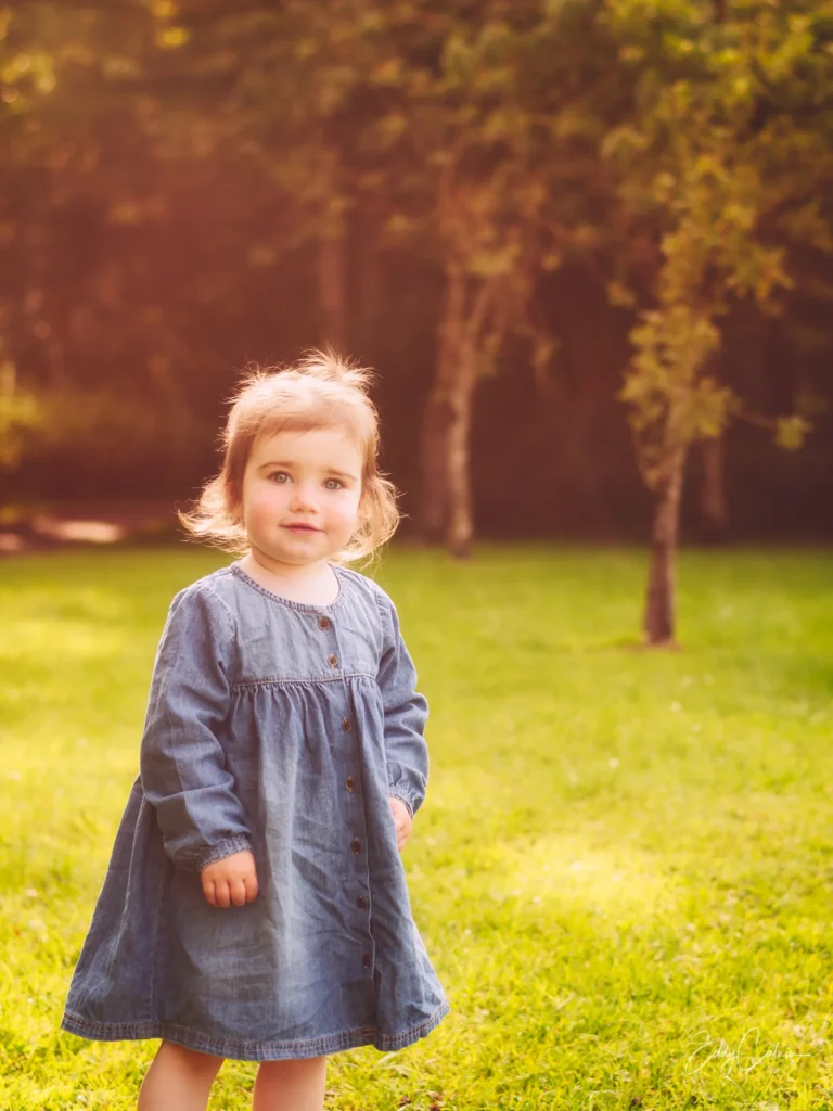 A young child wearing a blue denim dress stands on green grass with trees and sunlight in the background, perfectly capturing the kind of candid moment a family photographer cherishes.Family photographer