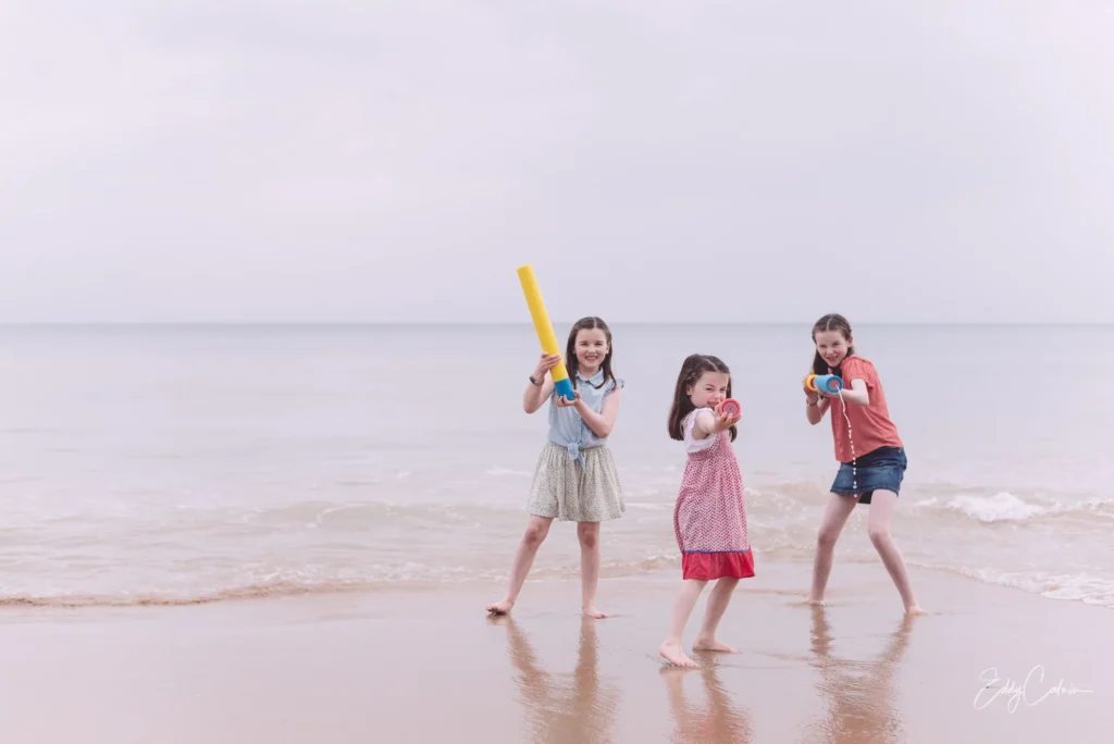 Three young girls stand at the edge of the beach, holding water toys and smiling whilst playing near the calm sea under a cloudy sky, perfectly capturing a candid moment any family photographer would cherish.Family photographer