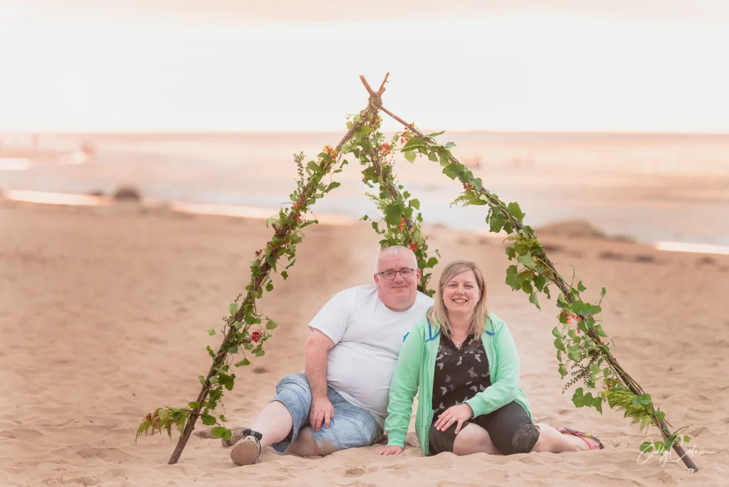 Two people sit on the sand under a simple wooden tepee frame decorated with greenery and flowers on a beach at sunset, perfectly capturing a heartfelt moment that any family photographer would cherish.Family photographer