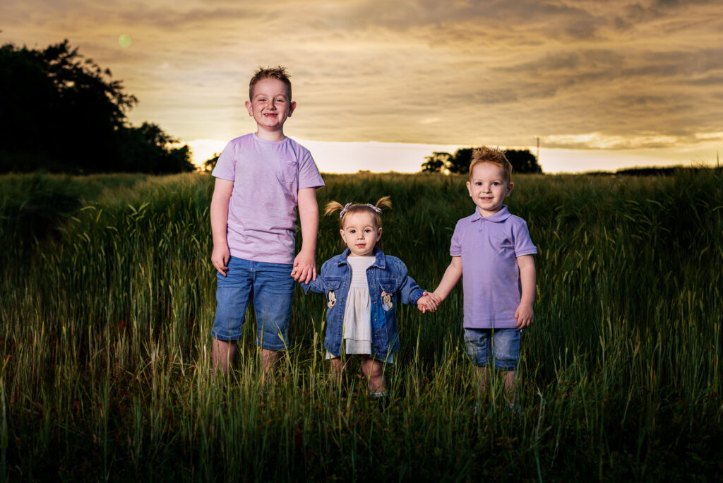 Northern Ireland outdoor family photographer capturing three smiling children holding hands in a golden field at sunset