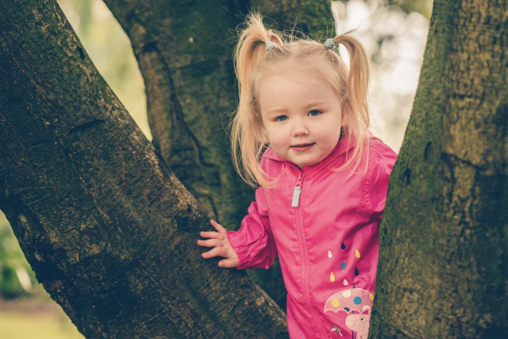 Northern Ireland outdoor family photographer portrait of a toddler in a tree wearing a pink jacket, playful natural session