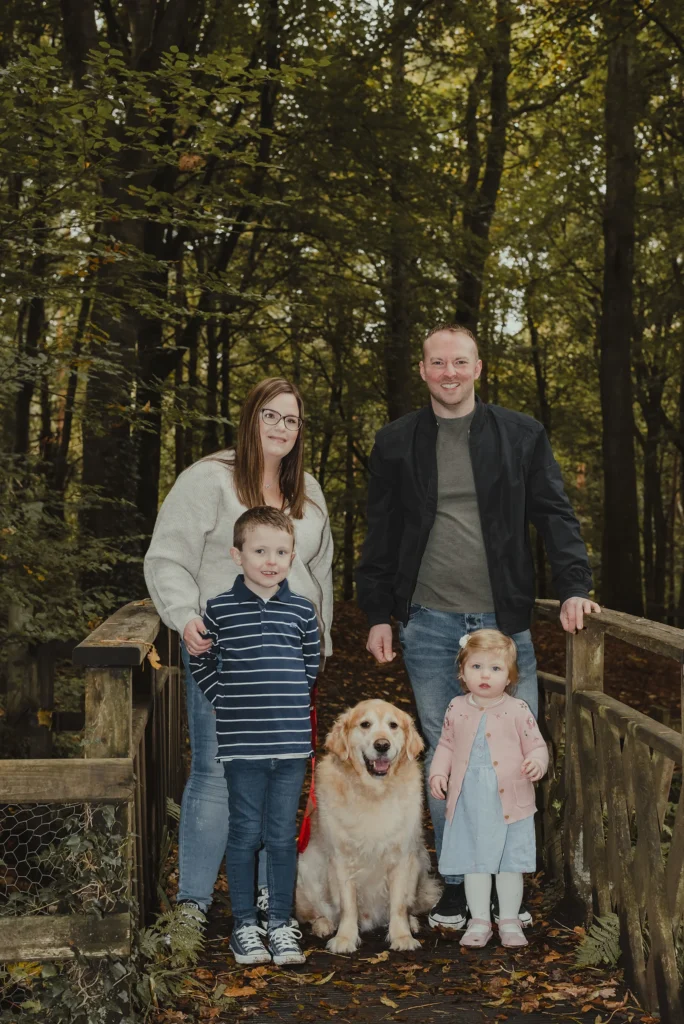 Northern Ireland outdoor family portrait on woodland bridge showing smiling parents, two children and a golden retriever dog