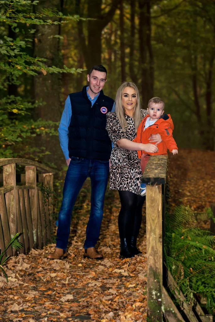 Family photographer in Northern Ireland captures parents and toddler on a woodland bridge surrounded by autumn leaves